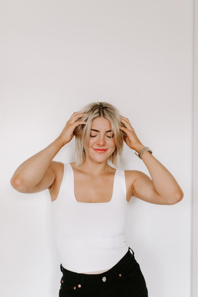 Courtney Johnson in a white tank top, hands running through her hair, eyes closed, against a white wall