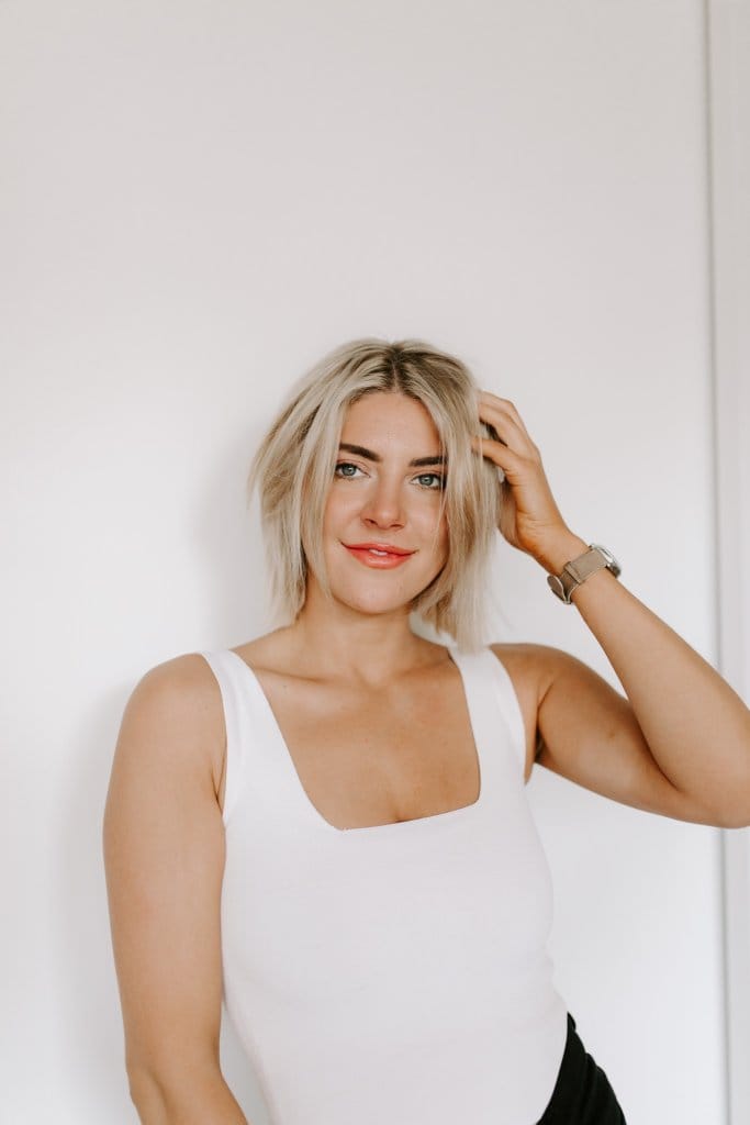 Courtney Johnson in a white tank top, one hand in her hair, smiling at the camera against a white wall
