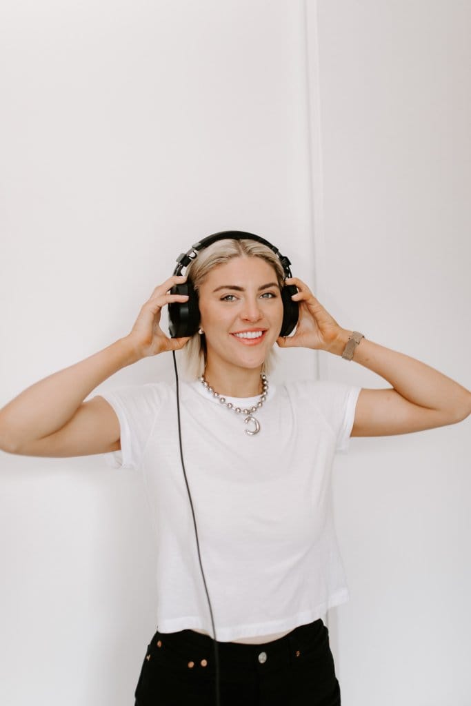 Courtney Johnson wearing wired headphones over a white t-shirt, smiling at the camera against a white wall