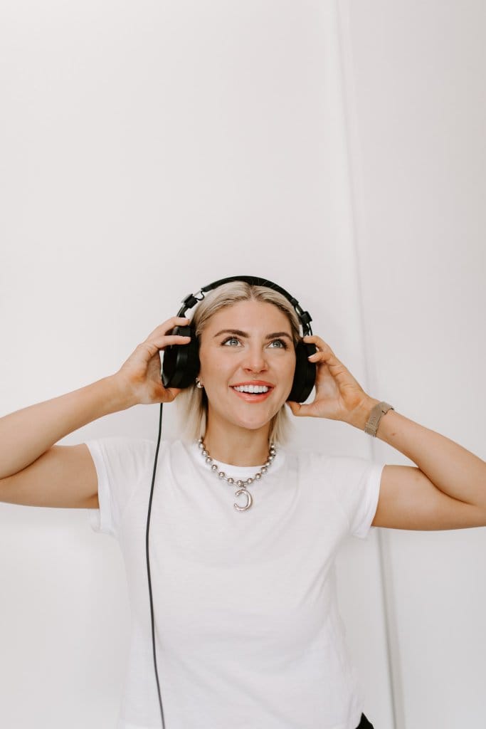 Courtney Johnson in a white t-shirt wearing wired headphones, looking up with an open smile against a white wall