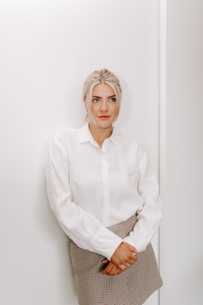 Courtney Johnson in a white button-down shirt and plaid skirt, leaning against a white wall
