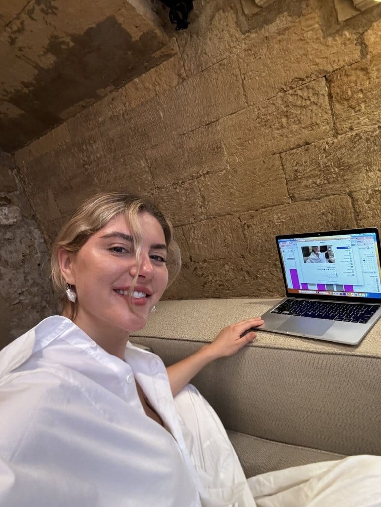 Courtney Johnson working on a MacBook at a stone-walled cafe, smiling at the camera